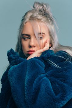 a woman with blonde hair and blue eyes is wearing a green coat, she has her hand on her chin