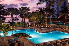 an outdoor swimming pool with lounge chairs and palm trees at dusk in front of the ocean