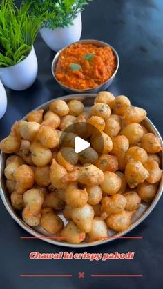 a bowl filled with potatoes next to two bowls of sauce and some green plants in the background