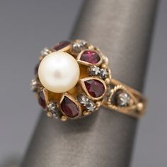 Crafted in mid-century with definite nods to Georgian design, this ring features a gorgeous glowing pearl atop bezel set rhodolite garnets separated by tiny diamond slices. The pearl is cultured and measures 7.41mm, with an ivory color and strong reflections. There are six of the rhodolite garnets- red with a pinkish hue. The garnets measure about 5 x 3mm, calculating to 0.15ct each. In between the bezels are tiny white gold floral designs holding round diamond slices. There is one on each side Georgian Ring, Gem Ring, Rhodolite Garnet, Tiny Diamond, Ivory Color, Red Garnet, Cultured Pearls, Round Diamonds, Garnet