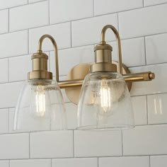 two clear glass shades on the wall light fixture in a white tiled bathroom with brass fixtures