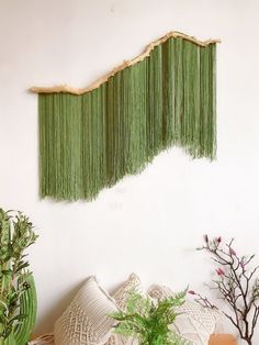 a green wall hanging on the side of a white wall next to a potted plant