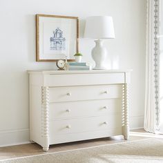 a white dresser with two lamps on top of it next to a painting and lamp shade