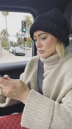 a woman sitting in the back seat of a car looking at her cell phone while wearing a beanie