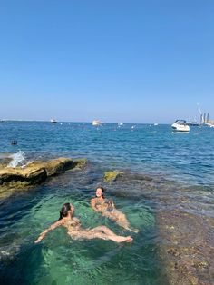 two people are swimming in the water near some rocks and boats on the ocean behind them