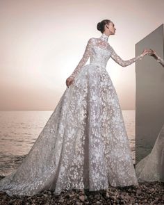 a woman in a wedding dress standing on rocks near the ocean with her hand on the wall