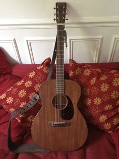 an acoustic guitar sitting on top of a bed next to a red pillow and pillows