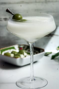 a martini glass filled with white wine and green olives next to a tray of food