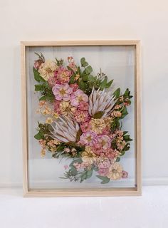 an arrangement of flowers in a wooden frame on a white surface with a wall behind it