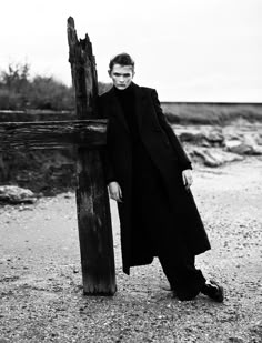 black and white photograph of a man leaning against a wooden cross on the beach with his hands in his pockets