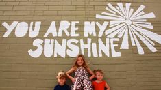 three children standing in front of a wall with the words you are my sunshine painted on it