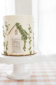 a white cake decorated with green leaves and a house on top is sitting on a pedestal