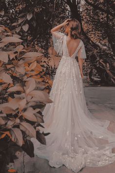 a woman in a wedding dress is standing near some bushes and trees with her back to the camera