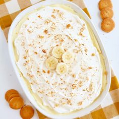 a banana pudding in a white dish on a checkered tablecloth with some cookies