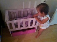 a baby standing in front of a white crib with pink bins on the floor