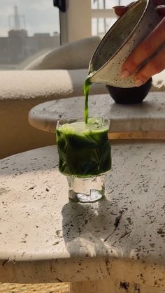 someone pours green liquid into a glass on a marble table top with a view of the city in the distance