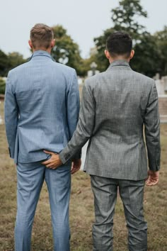 two men in suits standing next to each other on a grass covered field with trees behind them