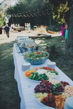 a table full of food and plates on it with people in the background at an outdoor event