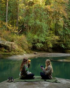 two young women sitting on the edge of a river drinking tea and looking at their cell phones