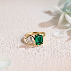 an emerald and diamond ring sitting on a white surface with flowers in the foreground