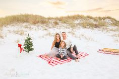 a family sitting on a blanket in the sand with a christmas tree and presents behind them