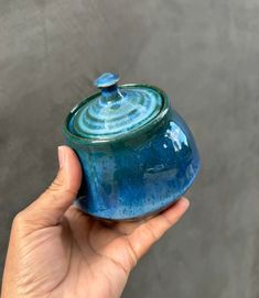 a hand holding a blue ceramic container with a lid in front of a gray wall