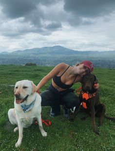 a woman kneeling down next to two dogs on top of a grass covered field with mountains in the background