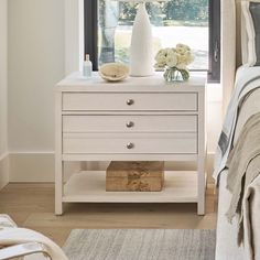 a white nightstand with flowers on it in front of a window and a bed next to it