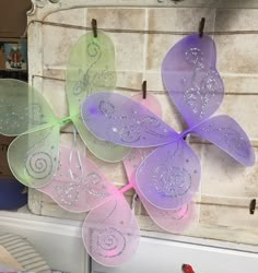 three purple and green butterfly decorations hanging from clothes pins on a white brick wall in front of a mirror