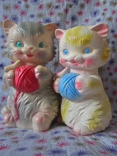 two ceramic cats sitting next to each other on a bed with blue eyes and one has a ball of yarn in its paws