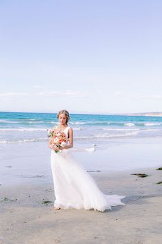 a woman standing on top of a sandy beach next to the ocean holding a bouquet