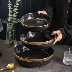 a person holding three black and gold bowls on top of a table next to a plant