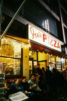 many people are sitting outside of a pizza shop at night, with lights on the windows