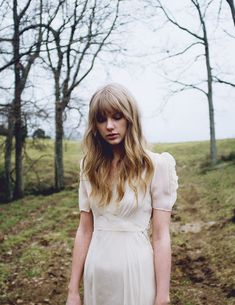 a woman in a white dress standing on a dirt road next to trees and grass