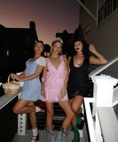 three women dressed in costumes posing for a photo on the balcony at night with one woman holding a basket