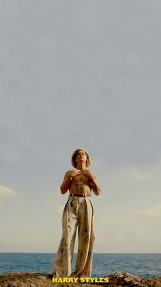 a woman standing on top of a rocky cliff next to the ocean with her hands in her pockets