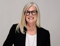 a woman wearing glasses and a black blazer smiles at the camera while standing in front of a gray background