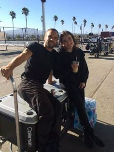 a man and woman sitting on top of a cooler in the middle of a parking lot