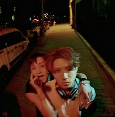 two young men standing next to each other on a sidewalk in the dark with their arms around one another