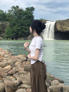 a woman standing in front of a waterfall with her hands up to her chest and looking at the water