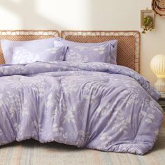 a bed with purple comforter and pillows in a white room next to a window