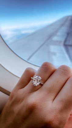 a woman's hand holding an engagement ring in front of the window of an airplane