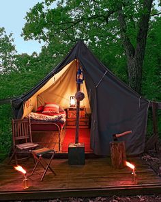 a tent is lit up in the woods with two chairs and a bed on it