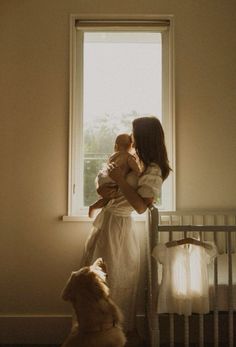 a woman holding a baby standing next to a crib with a dog looking out the window