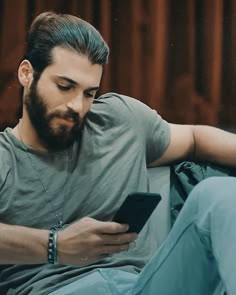 a bearded man sitting on a couch looking at his cell phone while wearing headphones