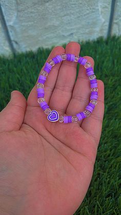 a hand holding a purple beaded bracelet with a heart on it in the grass