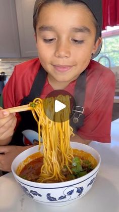 a young boy eating noodles with chopsticks in a bowl on the table next to him