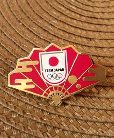 a red and gold pin with the team japan logo on it sitting on a woven surface