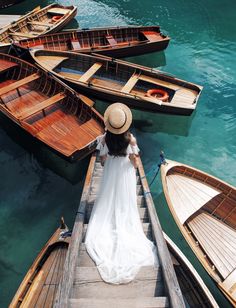 a woman in a white dress and straw hat standing on a dock next to row boats