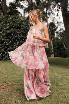 a woman standing in the grass wearing a pink floral print dress and holding a drink